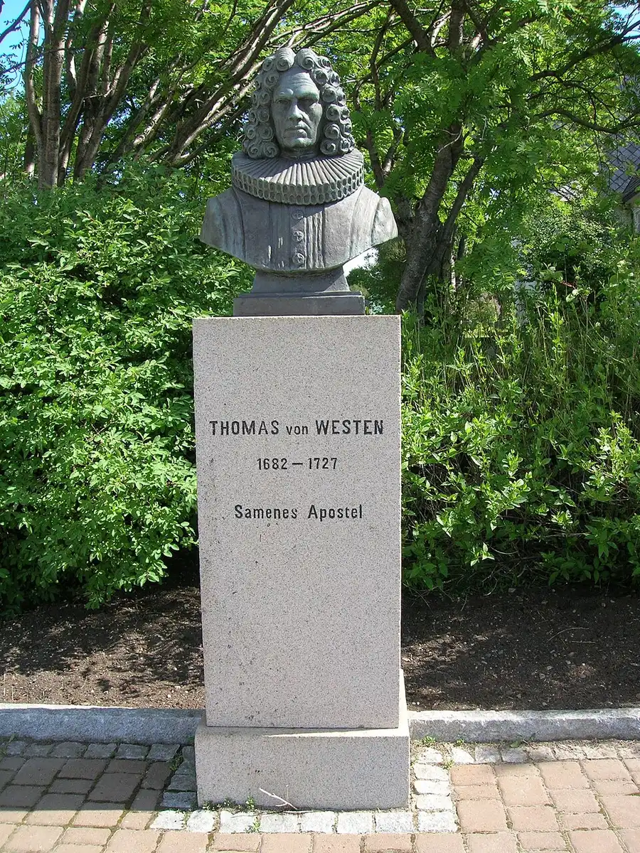 A weathered bronze bust of a bewigged man on a granite plinth inscribed "THOMAS von WESTEN 1682-1727, Samenes Apostel," set against greenery.