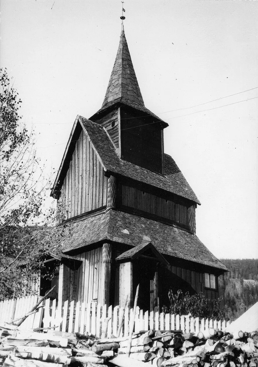 Black-and-white Riksantikvaren photograph of Torpo stave church from the southwest: tiered shingled roofs stepping up to the pyramid-capped turret, vertical staves and an entrance porch behind a white picket fence, stacked firewood in the foreground.