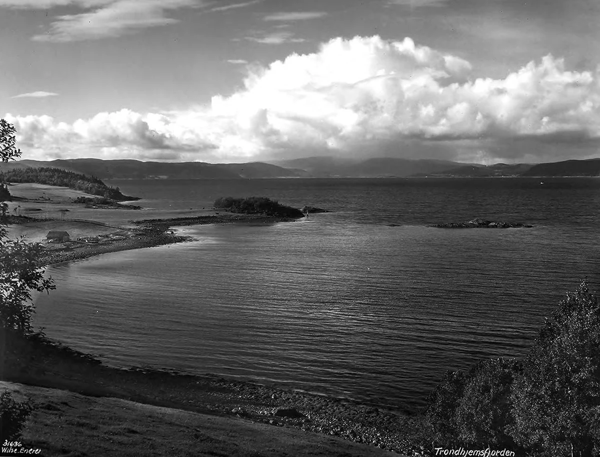 The Trondheim Fjord in Anders Beer Wilse's 1927 photograph — a curved stony shore with farm fields and a shed at the water's edge, a wooded islet, and the broad fjord running to distant hills under a towering cloud; "Trondhjemsfjorden" is inscribed in the negative.