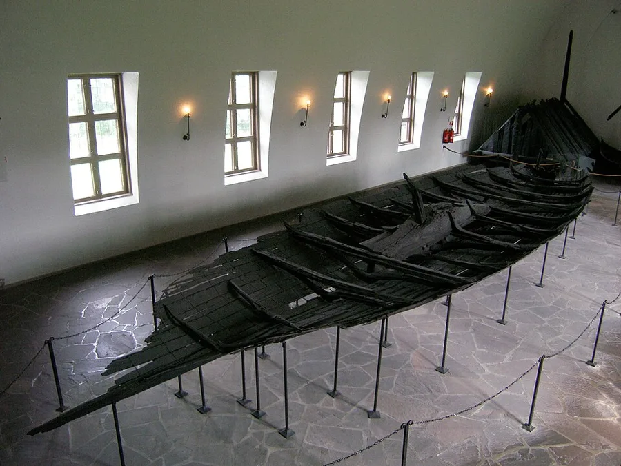 The fragmentary blackened hull of the Tune ship raised on slender metal posts in a white vaulted museum hall, broken strakes and exposed ribs lit by a row of slanted windows and wall lamps, above a flagstone floor edged with a chain barrier.