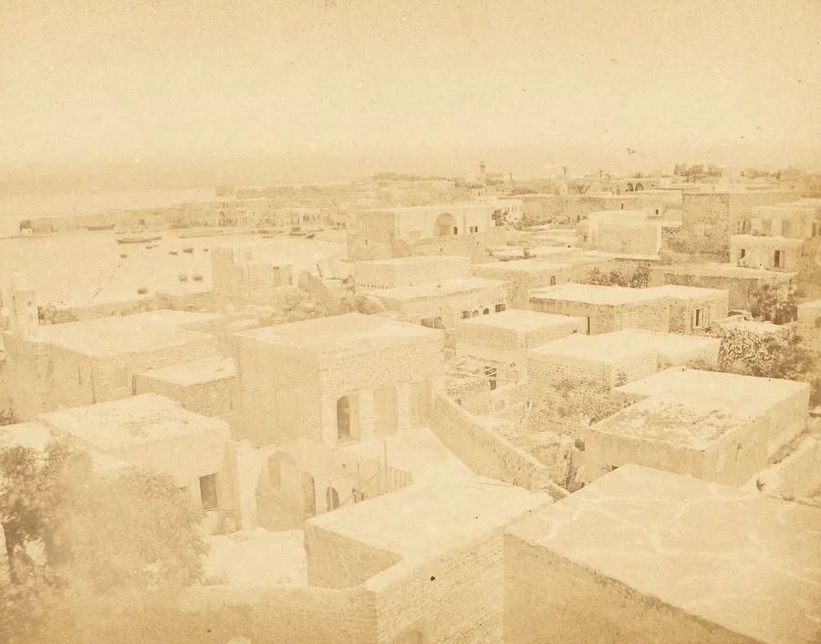 Sepia albumen photograph over the flat stone rooftops of Tyre, minarets rising among the houses, small boats moored in the curved harbor at left and the open Mediterranean beyond.