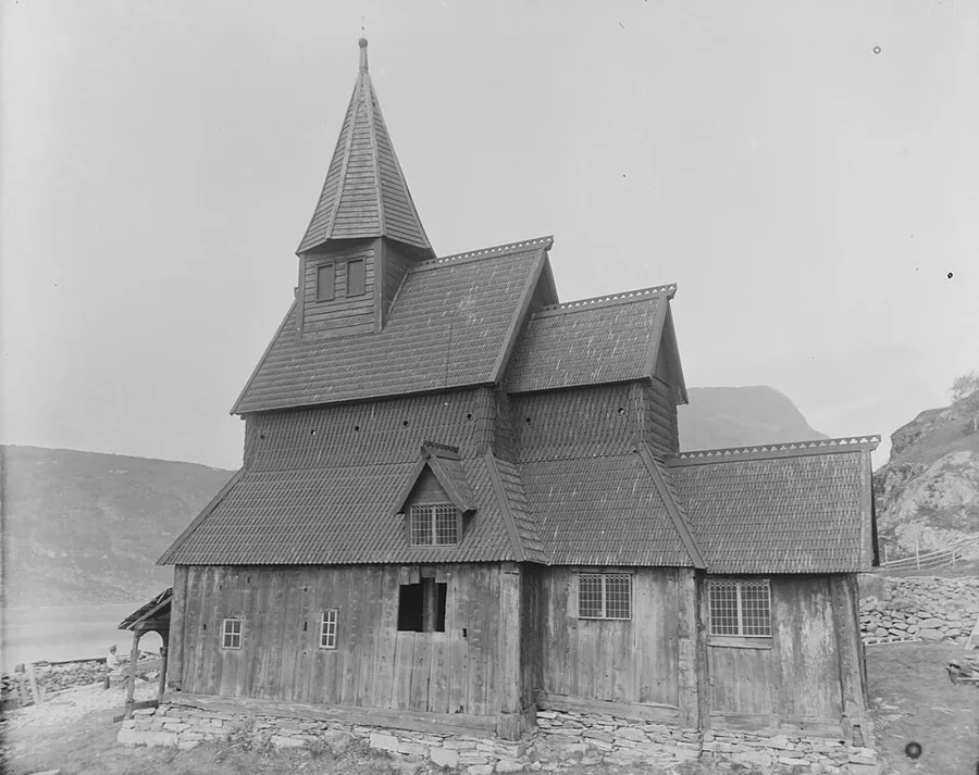 Late-1800s photograph of Urnes stave church from the southwest: the dark tiered shingle roofs and small turret rise over plank walls above the churchyard's stone wall, with the Lustrafjord and mountainside behind.