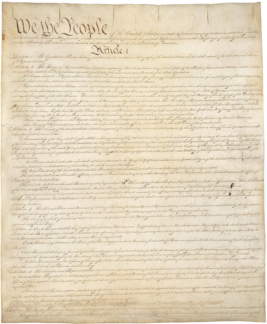 The engrossed parchment first page of the US Constitution, aged golden-brown, opening in large calligraphic script 'We the People' followed by dense handwritten Article I text in ruled columns.