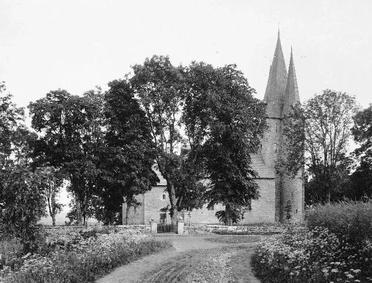 Photograph from about 1900 of Husaby church in Västergötland — its broad stone tower and twin spires rising over churchyard trees, with flowering meadows and a dirt lane in the foreground.