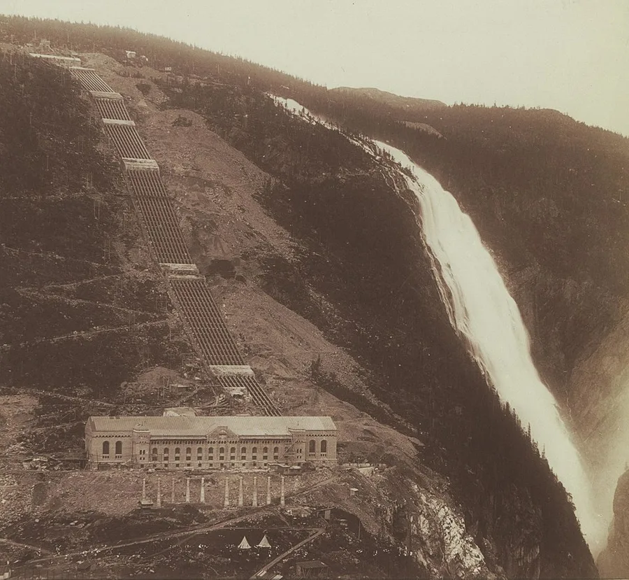 Sepia archival photograph of the Vemork power station, a long stone industrial building at the base of a steep mountainside, with rows of penstock pipes descending the slope above it and a tall waterfall cascading down the rock face beside it.