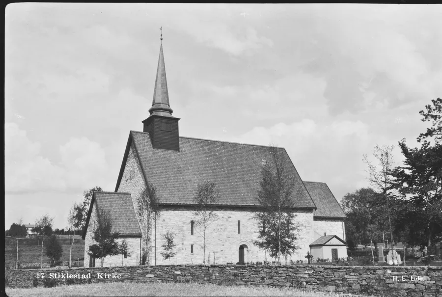 Early-20th-century photograph by H. E. Eide: Stiklestad church, a long stone building with a steep shingled roof and slender spire, stands behind its churchyard wall among young birches, with the low farmland of the Verdal valley opening at left.