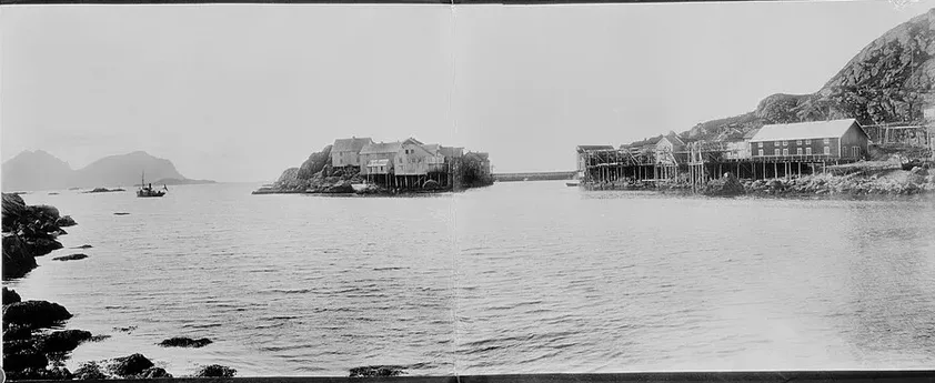 A panoramic harbour-survey photograph of Nyksund in Vesterålen from around 1900 — fishing-village buildings and drying racks on the rocky shore of the open coast.