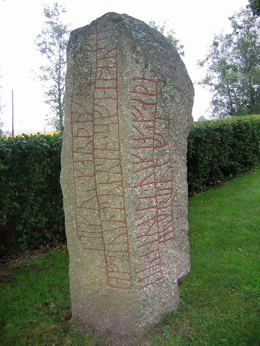 Color photograph of the Kälvesten runestone (Ög 8): a tall grey granite stone with runic inscription bands picked out in red running down two adjoining faces, standing on grass in front of a clipped hedge with trees behind.