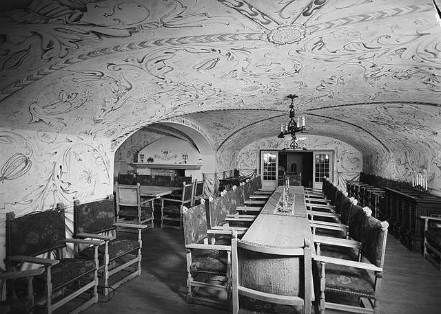 An ornate interior hall in Villa Grande — Quisling’s wartime residence ‘Gimle’ at Bygdøy, Oslo: a vaulted ceiling painted with dense scrollwork above a long banquet table flanked by carved high-backed chairs.