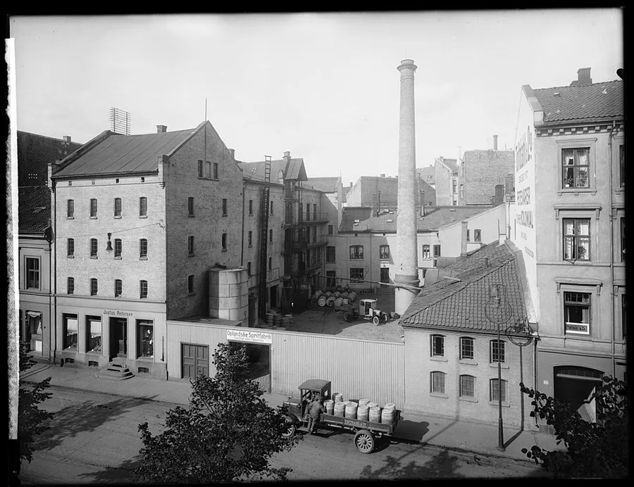 A 1925 photograph from an elevated vantage over an Oslo building complex: a brick warehouse with a “Justus Pedersen” shopfront, a tall factory chimney, an “Oplandske Spritfabrik” loading bay stacked with barrels, and a truck being loaded at the curb.