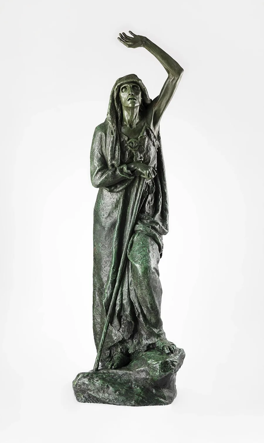 A weathered bronze statue, green with verdigris, of a hooded, cloaked woman standing on a rock, one arm raised high overhead with fingers splayed, the other hand resting on a long staff — photographed against a plain white background.