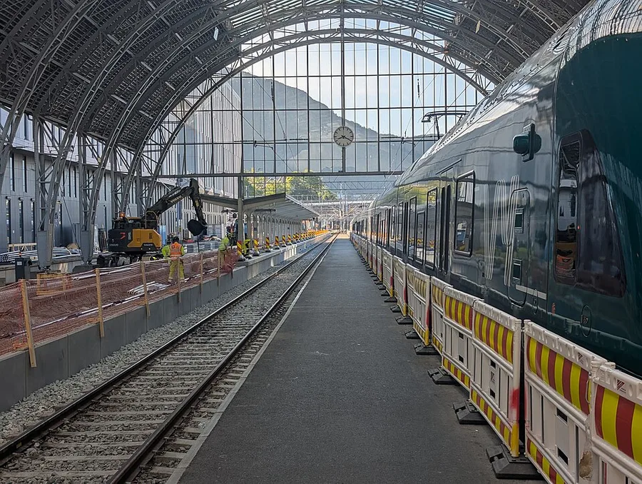 A dark green-and-white Vy passenger train at a platform under Bergen station's arched glass-and-iron roof, with track work, an excavator, and yellow safety barriers on an adjacent line under renovation.