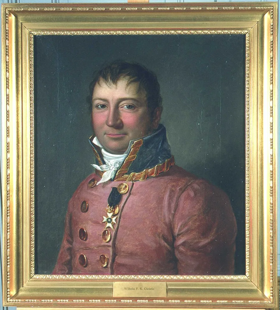 Framed oil portrait of a man with short reddish-brown hair in a rose-pink coat with a dark velvet collar trimmed in gold braid, gold buttons, a white cravat, and a white cross-form order on a ribbon; frame plate reads 'Wilhelm F. K. Christie.'