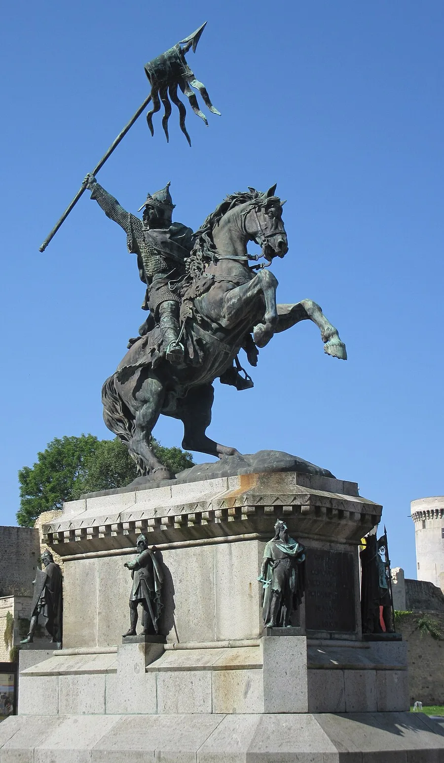 A bronze equestrian statue of William the Conqueror in mail hauberk and pointed helmet raising a long gonfanon banner from a rearing horse, atop a tall granite pedestal ringed with standing bronze dukes, against a clear blue sky with a castle tower behind.