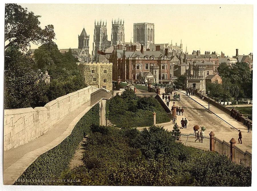 An 1890s colour photochrom labelled 'York from City Walls': the pale stone wall-walk curves through greenery in the foreground, past red-brick houses and a busy street with carts and pedestrians, toward York Minster's three great towers on the skyline.